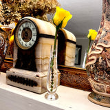 A vintage-style cream and brown marble mantel clock with a round clock face, displayed on a shelf next to a tall patterned ceramic vase and a small clear Waterford crystal bud vase holding a yellow rose.