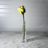 A single yellow rose bud in a Waterford crystal bud vase, placed on a white marble countertop against a white paneled wall.